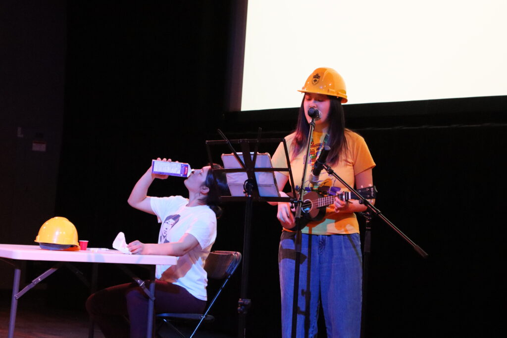 Two students performing a song about the CIV102 problem sets. One student in the foreground wears a hardhat and is playing a ukulele while singing. The second student is sat at a table with their hardhat resting on the table in front of them, they are drinking milk straight out of the carton a shot glass sits on the table in front of them.